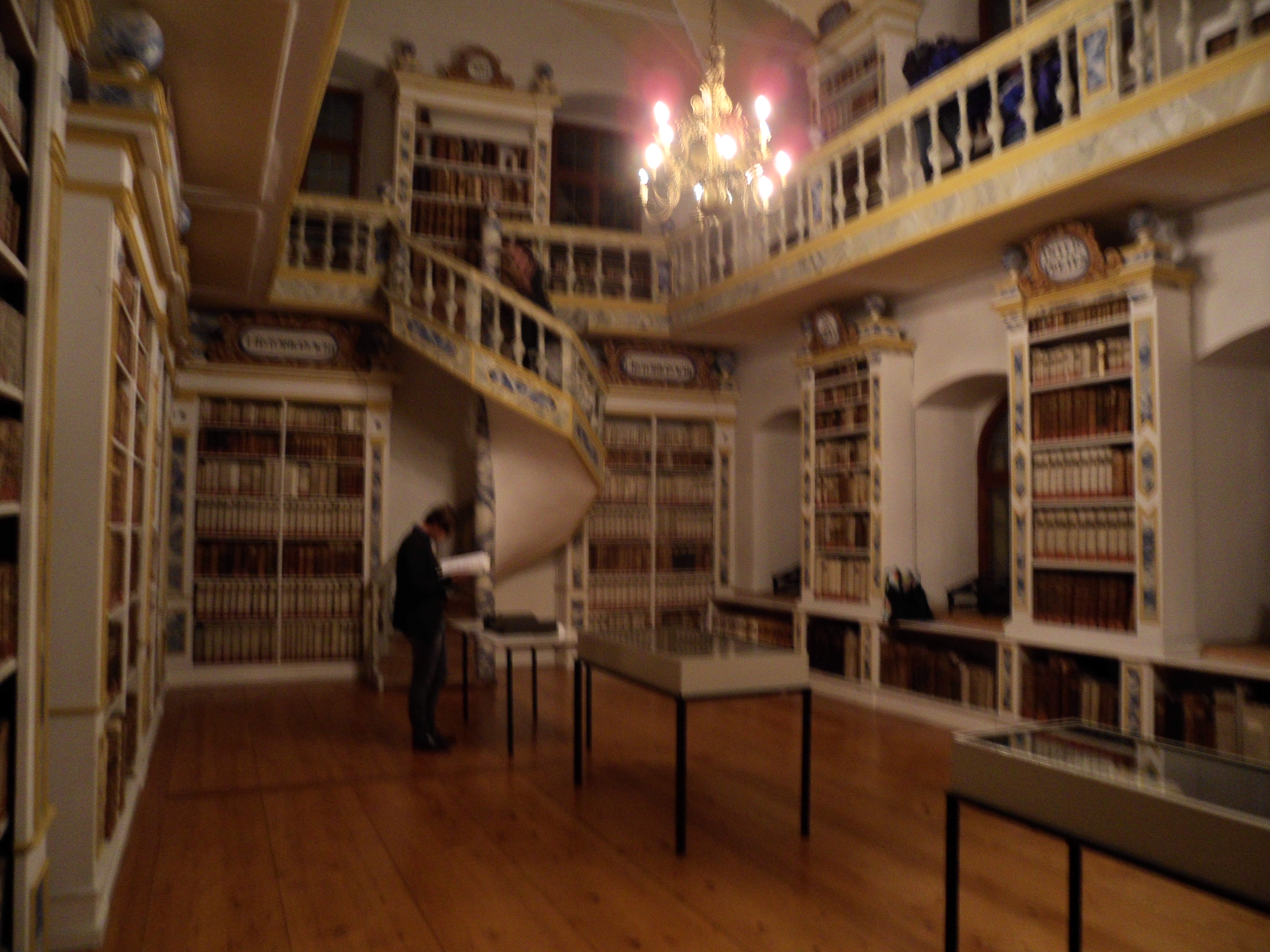 A large hall with glass windows, bookshelves between them, and a balcony encircling the room accessed by a spiral staircase
