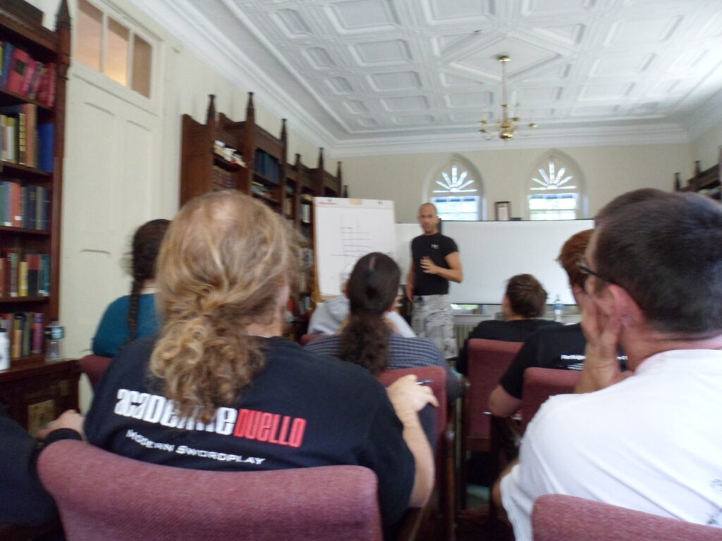 A library in a Neo-Gothic building with an athletic-looking man in shorts and a teeshirt talking in front of a portable whiteboard while an audience in chairs listens.