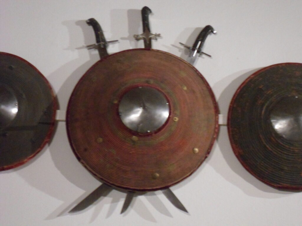 A round, domed wicker shield with a spiked steel boss and a cloth-bound rim. Three short scimilars hang behind it with their handles up and blades crossed at the middle.