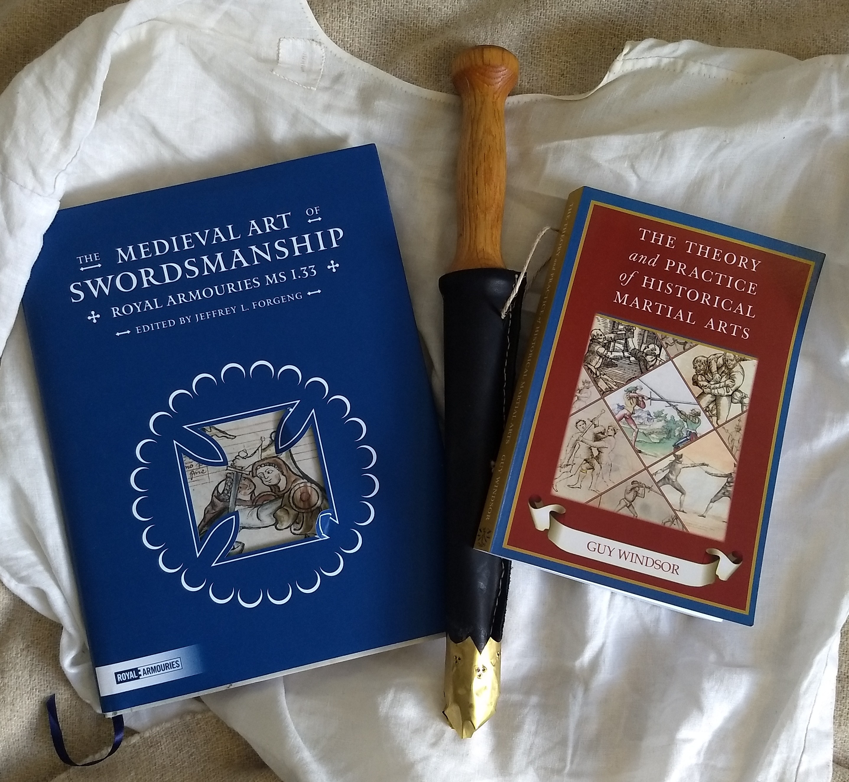 Two books with a wooden rondel dagger in a leather scabbard with a brass chape between them, laying on top of a hand-sewn linen shirt
