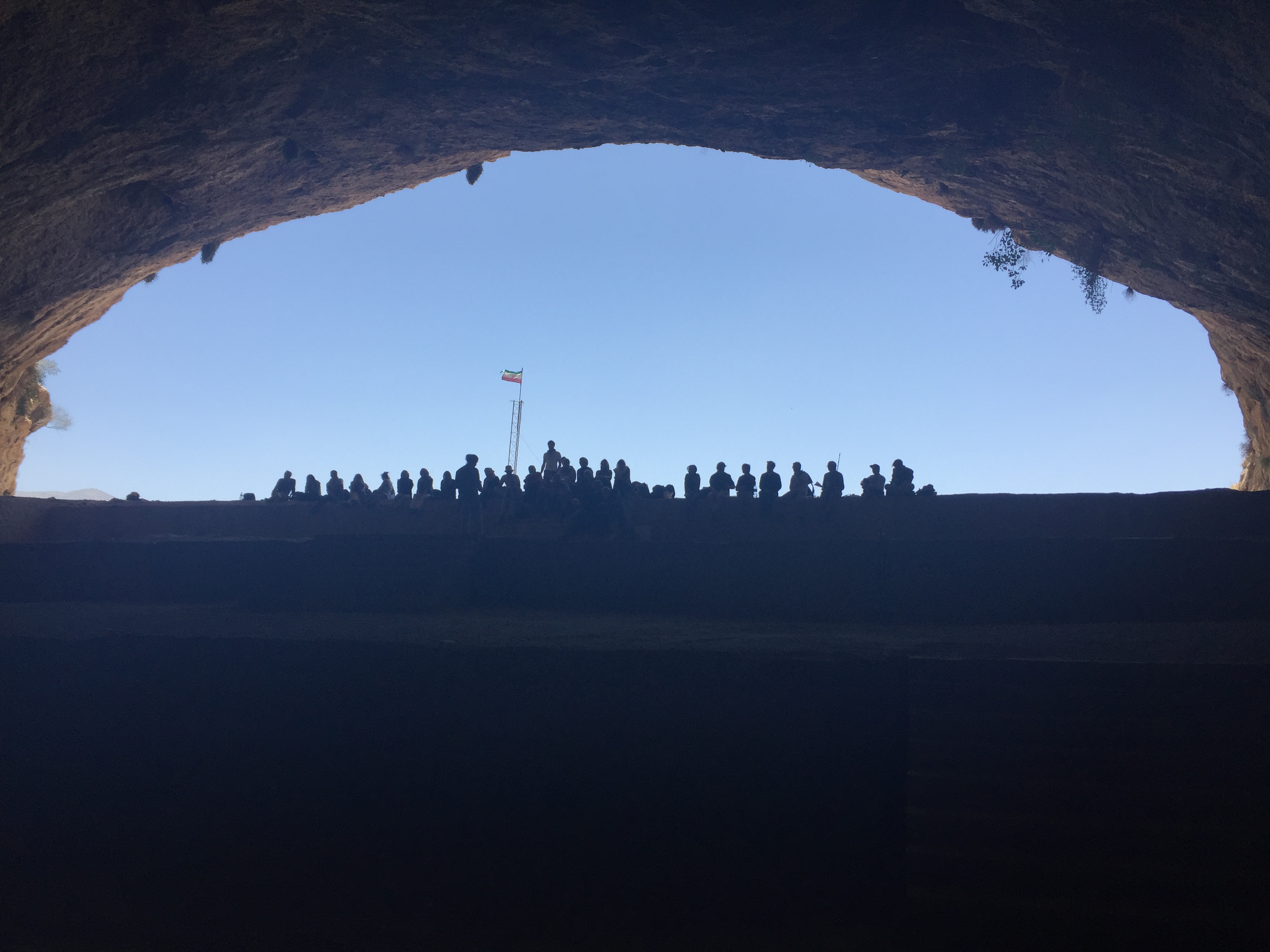 a group of seated people silhouetted against the sky of a cave mouth with an Iranian flag in the background