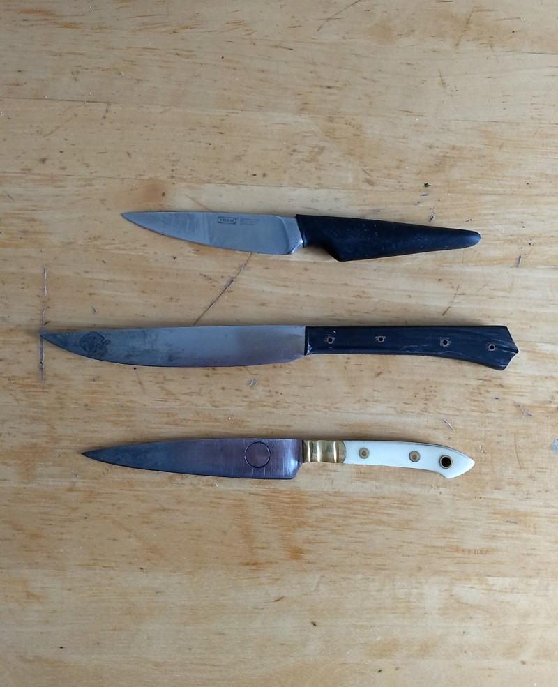 three paring knives (two hand-ground with steel blades and one mass-produced with a stainless steel blade) on a wooden table