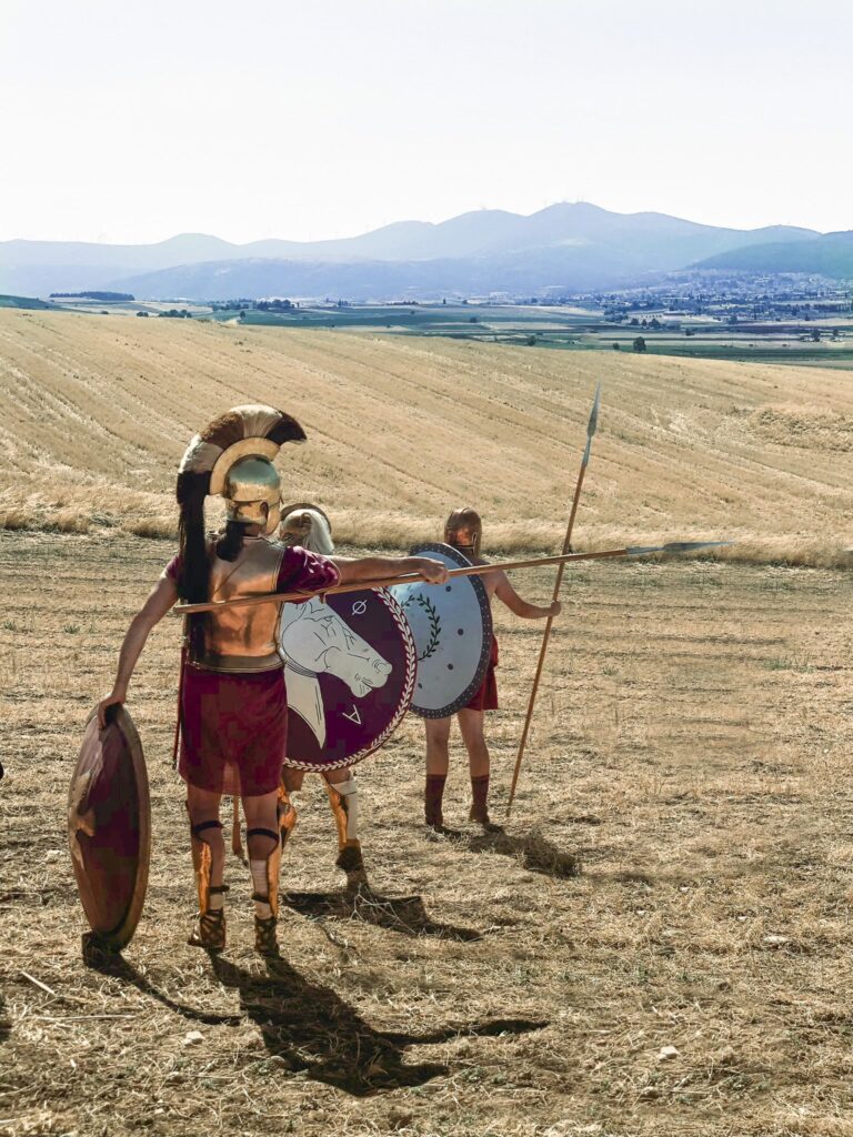 two men in hoplite kit stand on a dry field with a town in the background