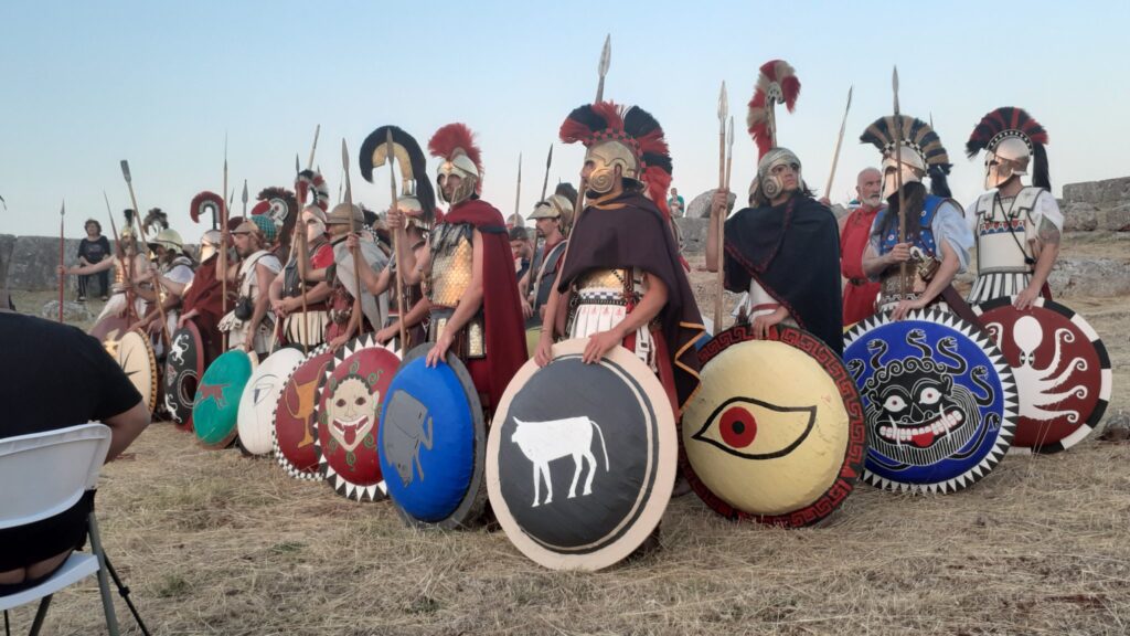 a group of hoplite reenactors formed in ranks and files next to a ruined wall