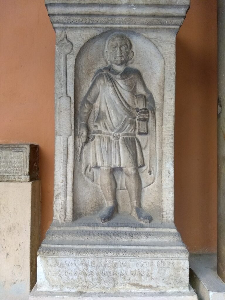 overview stele of the funerary stele of a Roman soldier. The stele is carved of coarse grey stone, probably limestone or similar. He stands in an arched niche wearing a cloak and tunic with a large belt. A spear with a bundle around the shaft is to his right, and he holds a scroll in his left hand.