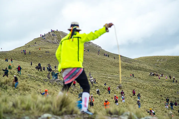 a man in a flourescent jacket over a Peruvian wrap brandishes a sling while warriors in the distance are silhouetted against a hill