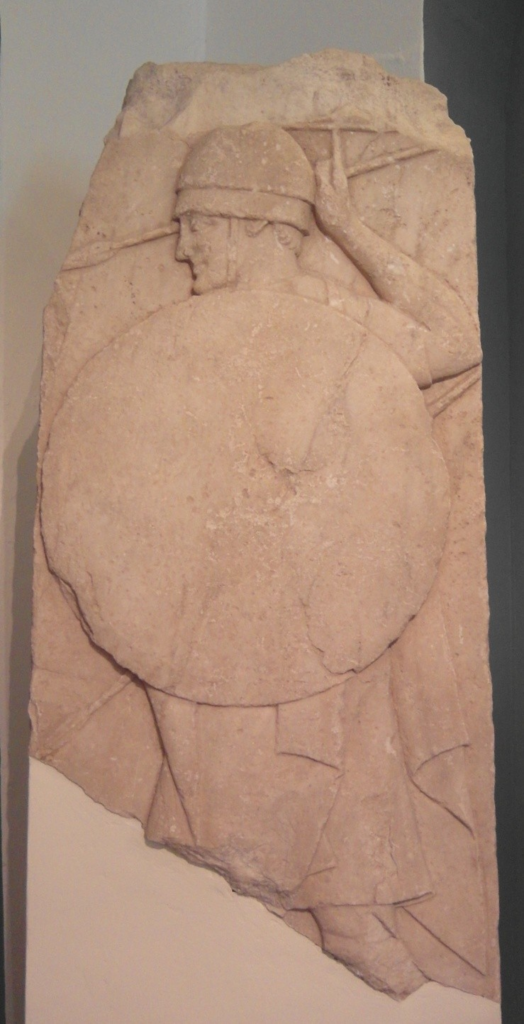 a colour photo of a grave stele showing a man with a helmet, a large round shield, and spears from his left hand side (ie. shield side)