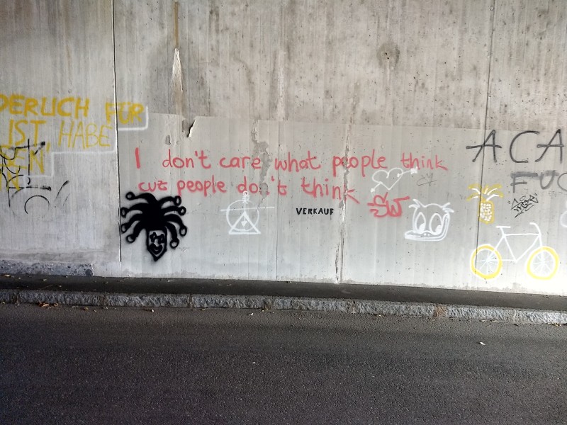 a red graffito on concrete: I don't care what people think cuz people don't think (nearby are other graffiti in black, white, and yellow)