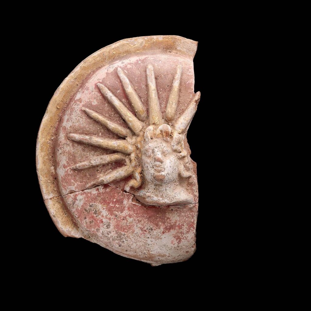a colour photo of a painted terracotta model of a Greek shield with the head of a god crowned with rays of light