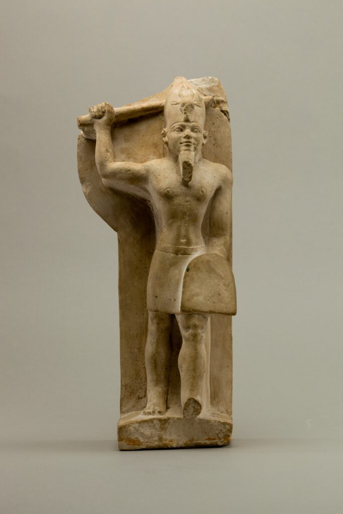 a plain linestone statue of a god with a tall hat on his head, a club or axe in his right hand, and a tombstone-shaped shield in his left hand