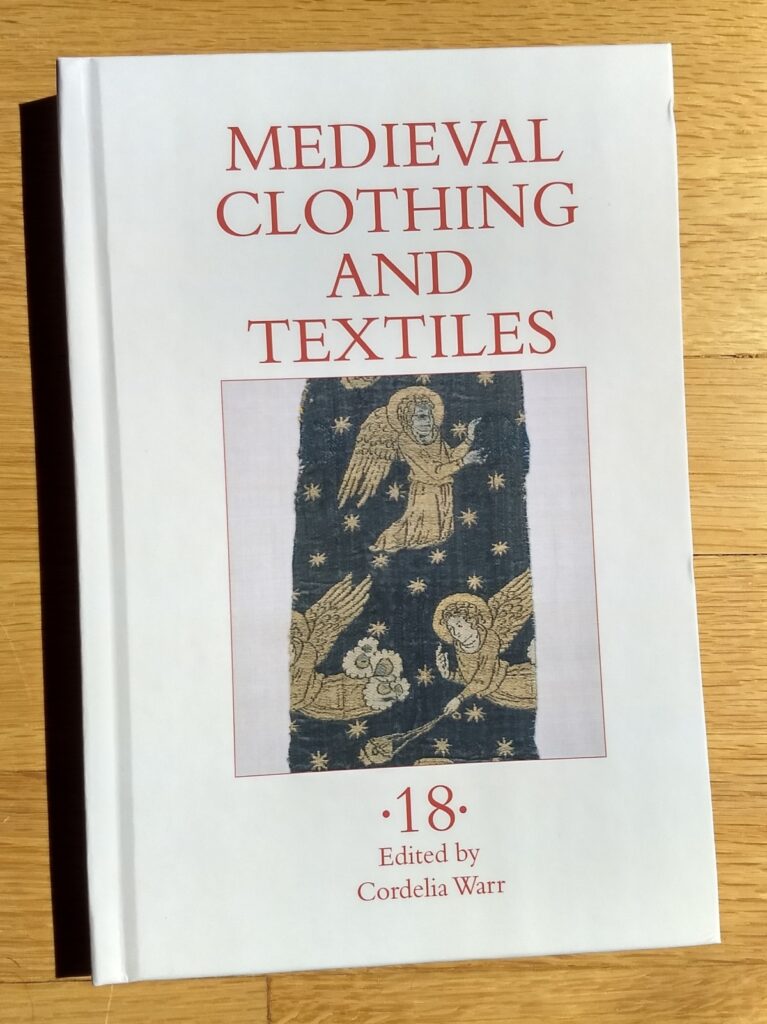 the cover of Medieval Clothing and Textiles 18, a thin hardcover book, in a patch of sunlight on a hardwood floor