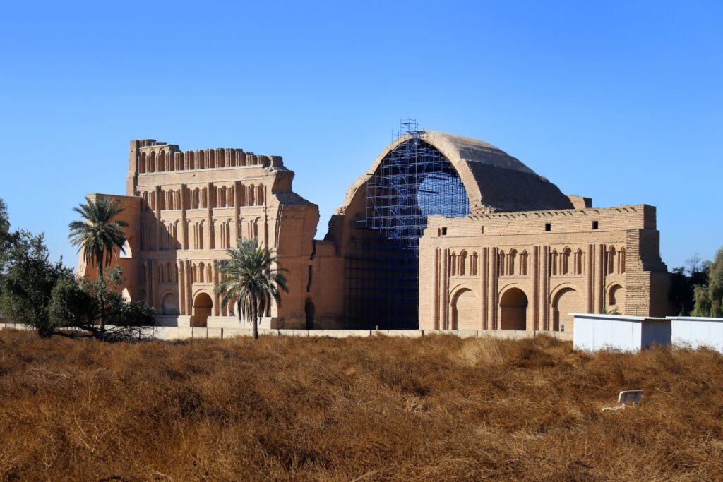 a colour photo of two lengths of baked-brick facade with fake arches and fake windows and a wide barrel-shaped brick arch in between. A scaffold holds up the front of the arch. Palm trees and dormant grass are in the foreground.