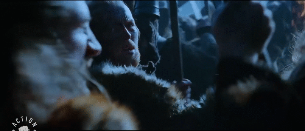 a black and blue screenshot of a man in furs and leathers looking back over his shoulder as ranks of men stand on either side holding shields and brandishing their weapons