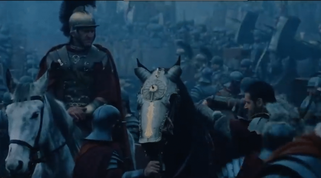 two Roman cavalry. One has a horse with a steel chaffron decorated with brass. The rest of the scene is dominated by blue, black, and brown tones.